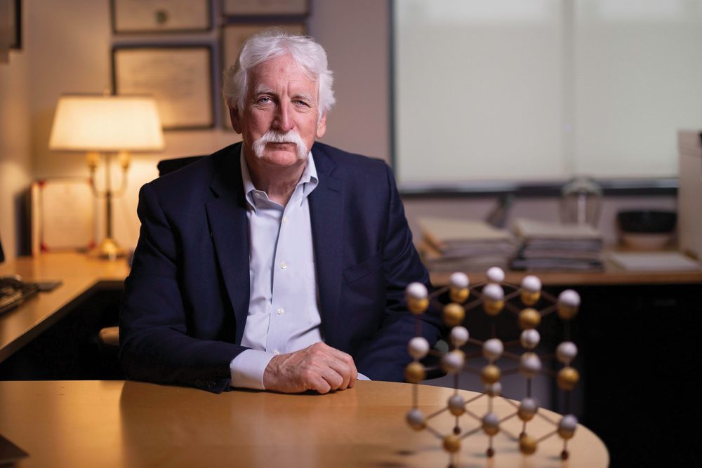 William L. Jorgensen seated at his desk beside a ball-and-stick molecular model, framed degrees on the wall behind him
